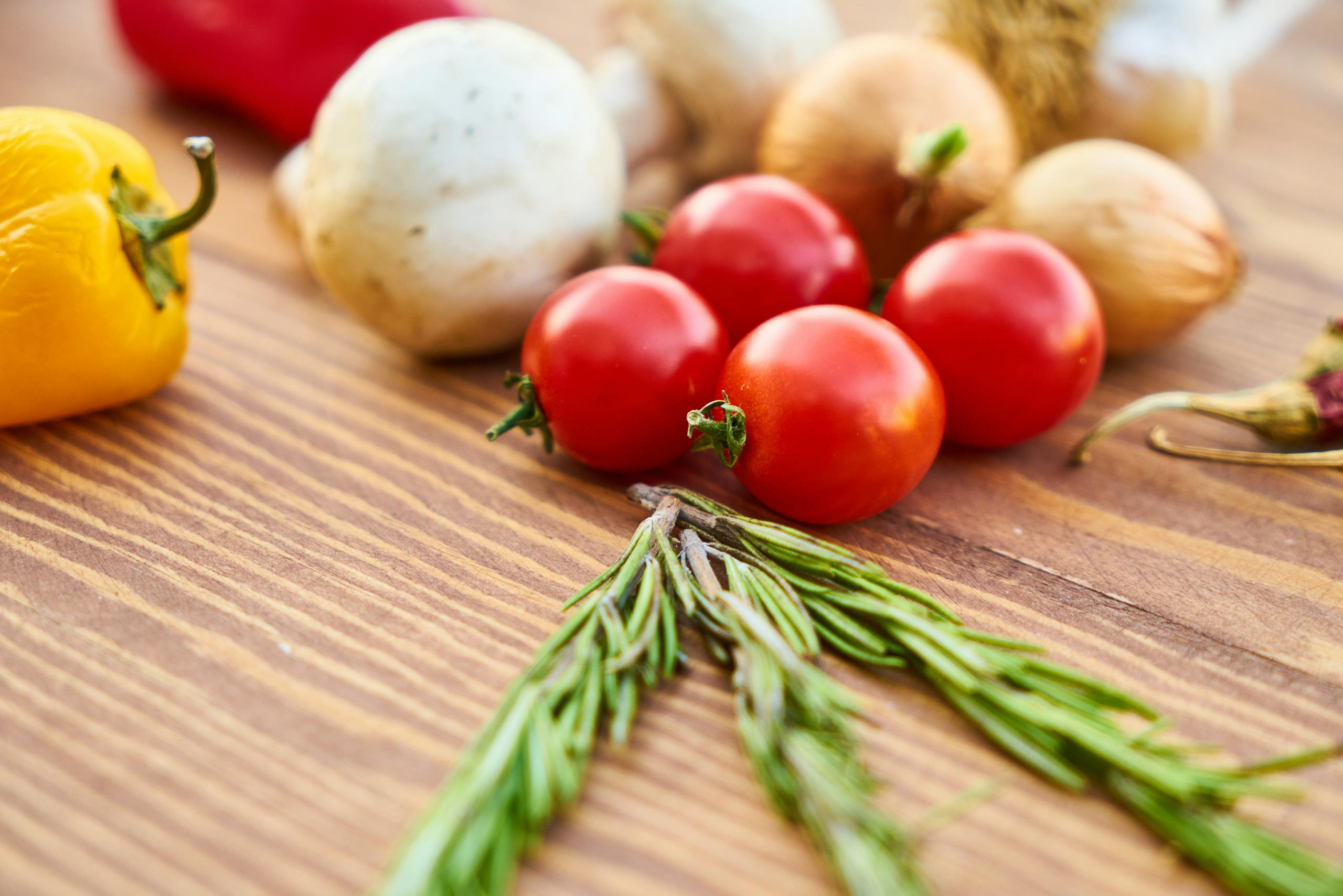 A variety of fresh vegetables including tomatoes, bell pepper, and rosemary on a wooden table.
