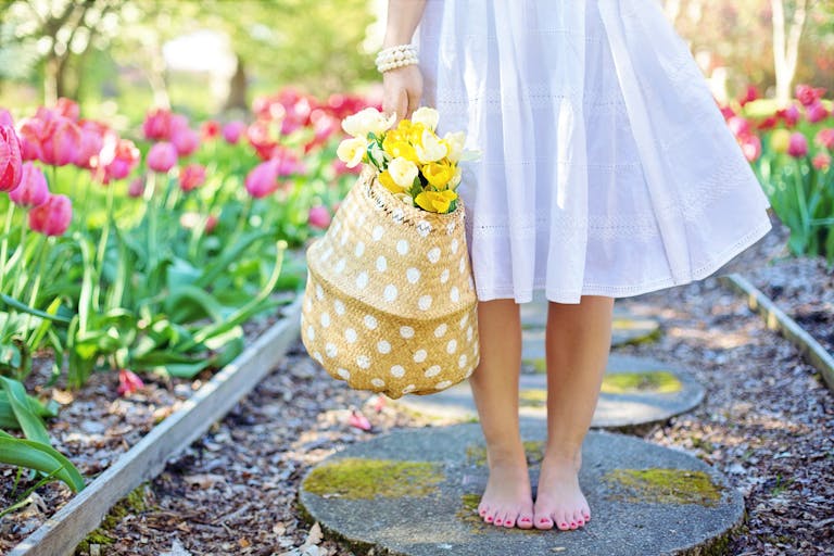 A woman in a white dress carries a basket of yellow flowers while walking barefoot in a tulip garden.